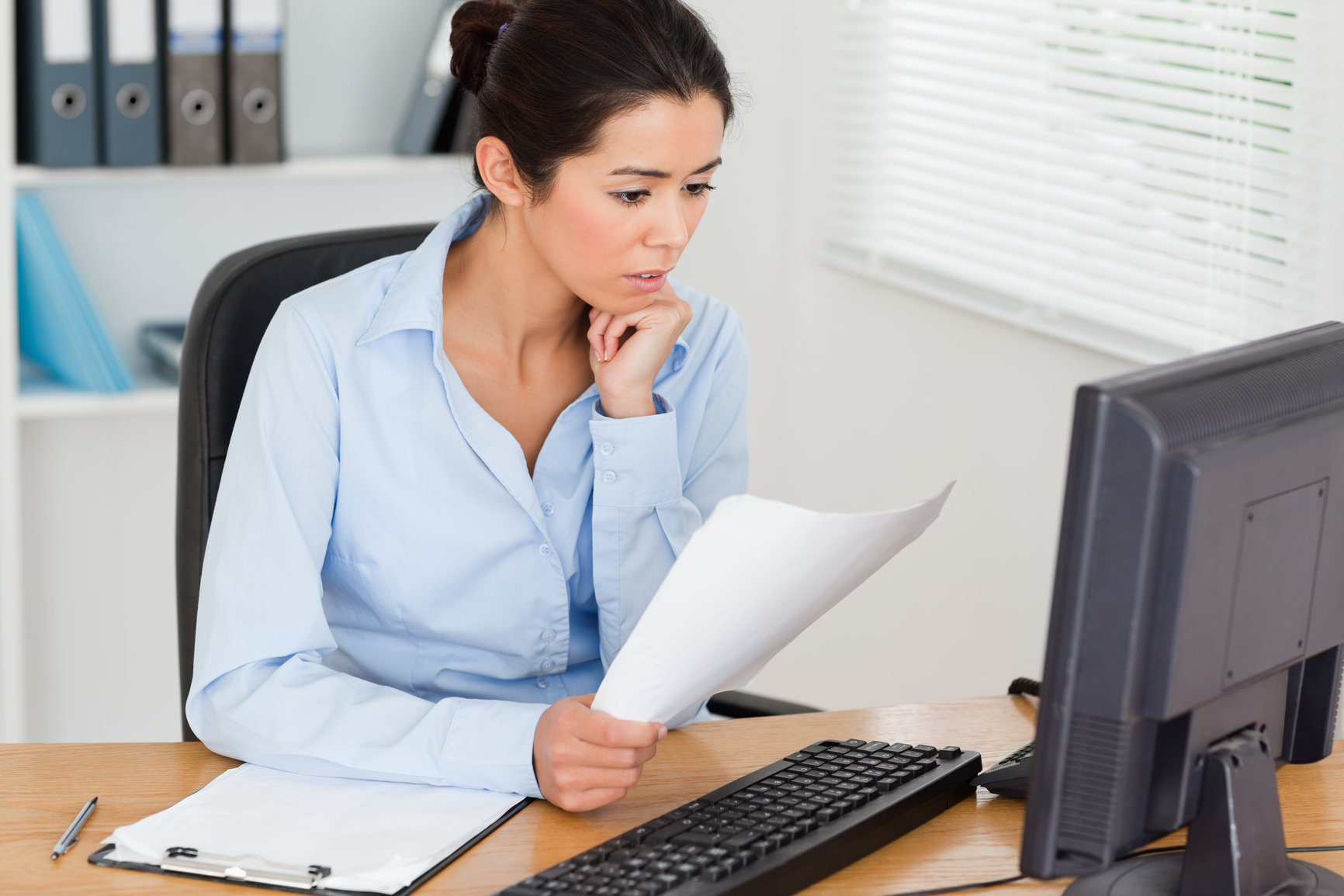 Gorgeous woman looking at a computer screen while holding a sheet of paper at the office